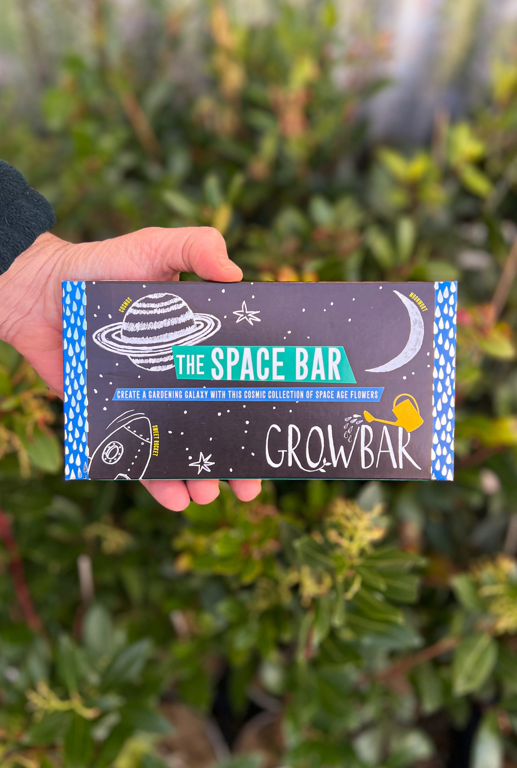 A beautiful decorated growbar package illustrated with a planet, sliver of a moon. stars and a space rocket held by a hand in front of green foliage.