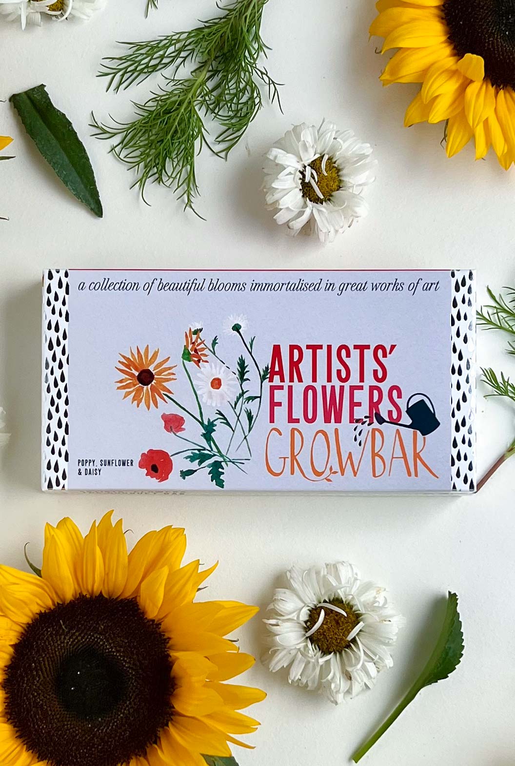 A grey Growbar package illustrated with colourful flowers. on a white table surrounded by two large yellow sunflowers, two white Daisy flowers and green leaves of Poppy flower plant.