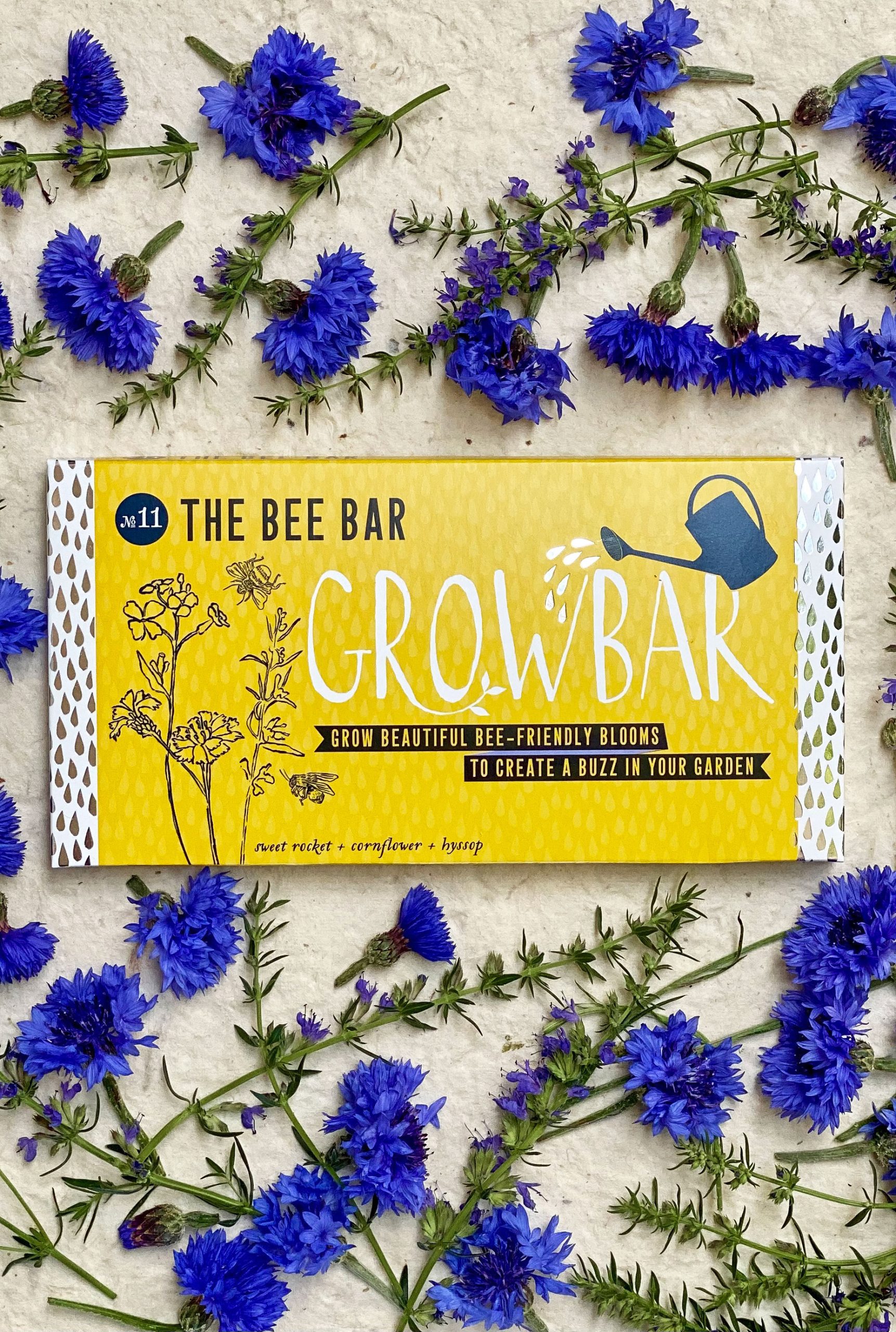 A yellow grow bar package surrounded by a decorative arrangement of bright blue Cornflowers and green twigs of Hyssop with small purple flowers.