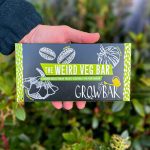 A black and green growbar for growing 'weird vegetables' held by a hand in front of green foliage.