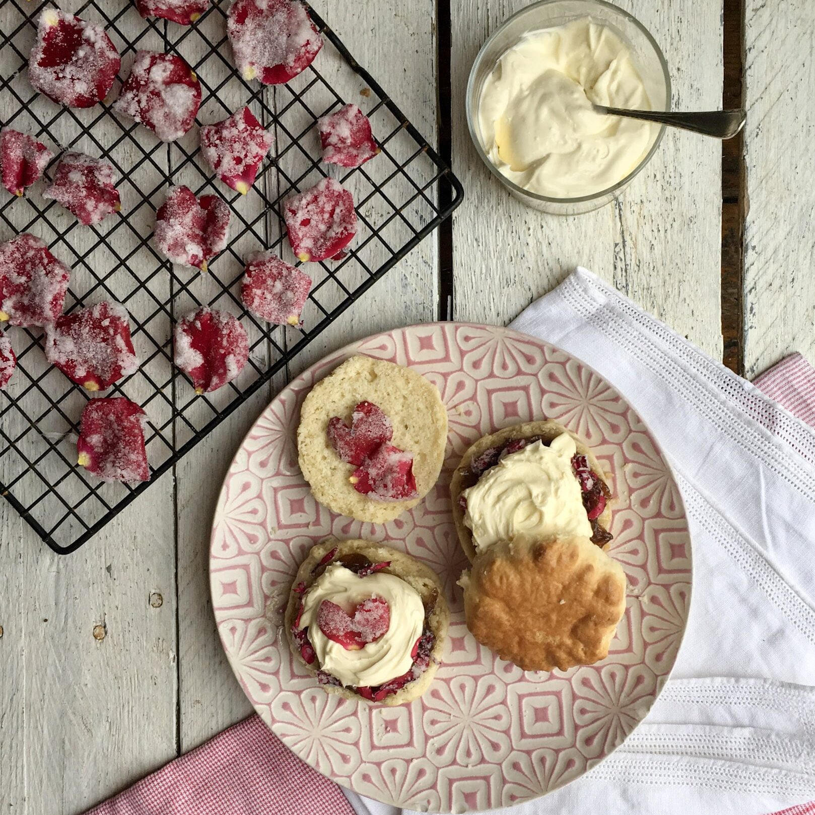 A wooden table set with a rose coloured plate with two scones, halved and spread with cream cheese, jam and candied rose petals, places alongside a white cotton napkin and a tray with sugared edible rose petals.